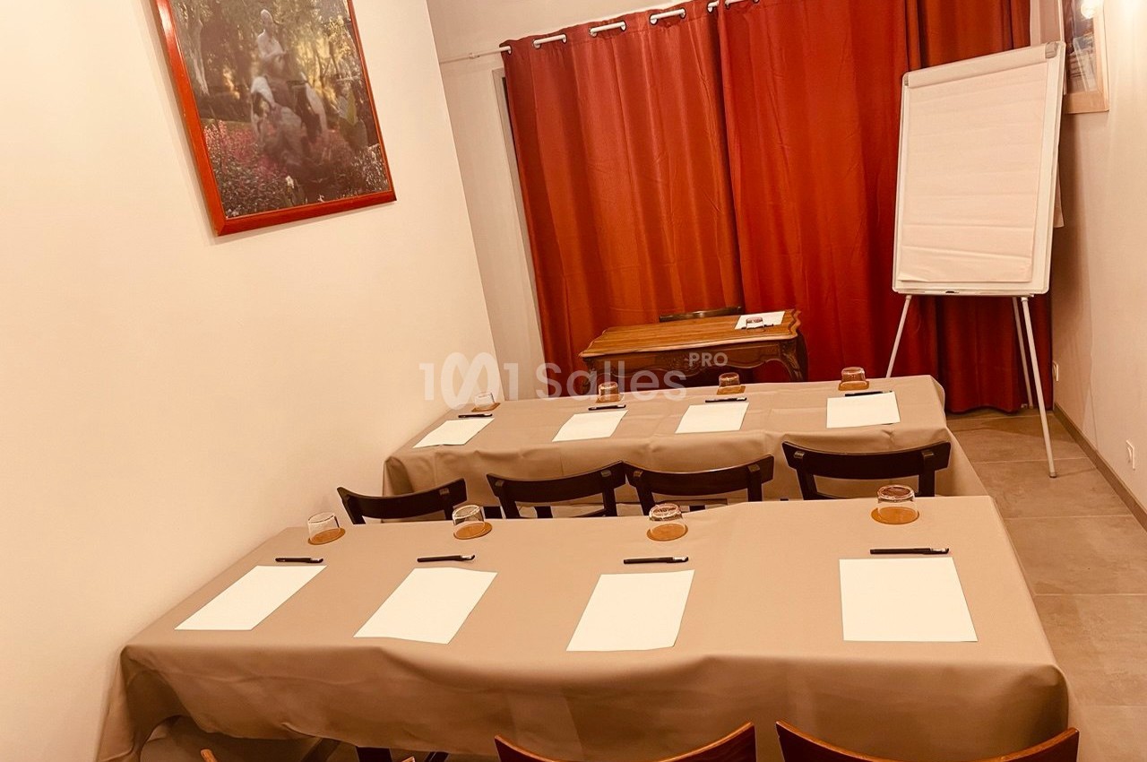 Salle de réunion avec tables disposées en rangées, blocs-notes et stylos, tableau blanc et rideaux rouges en arrière-plan.