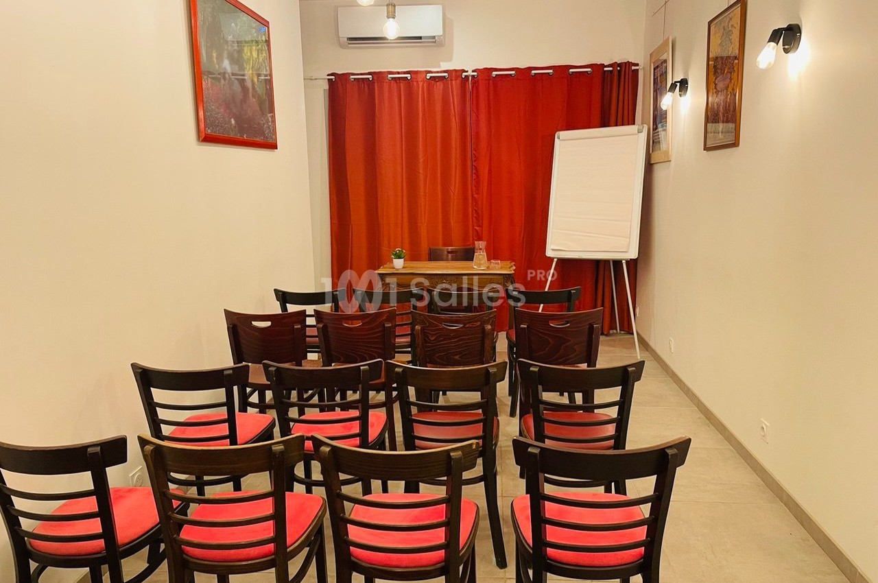 Salle de réunion avec chaises en bois, coussins rouges, rideaux rouges, tableau blanc et bureau en bois.