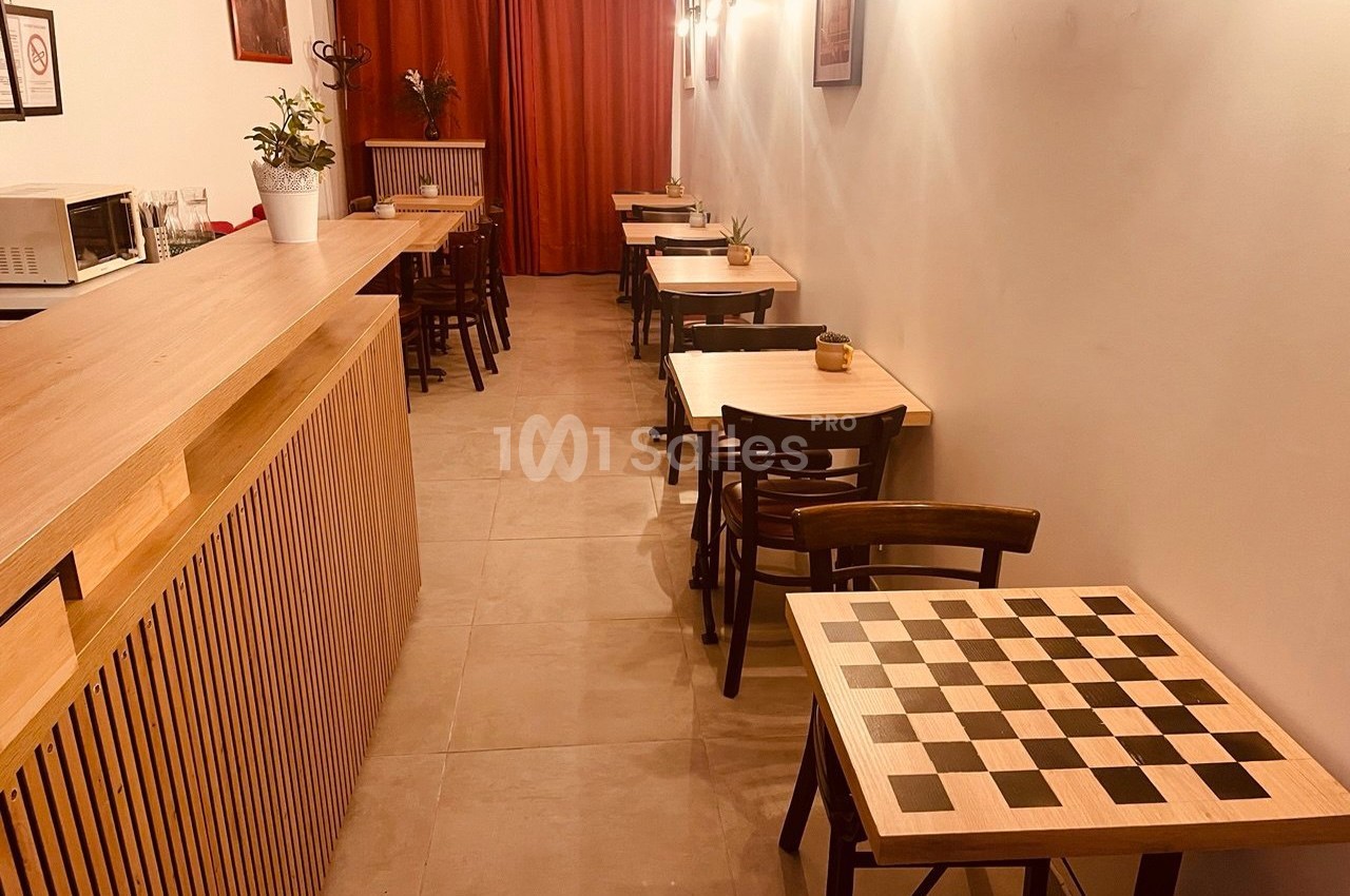 Salle de café avec tables en bois, tabourets, échiquier au premier plan et rideaux rouges en arrière-plan.