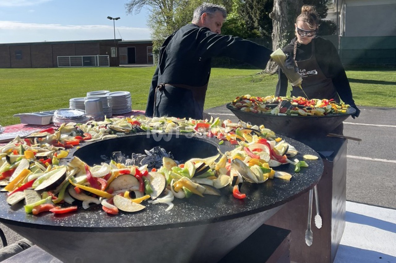 Deux personnes cuisinent des légumes variés sur de grandes plaques chauffantes en extérieur, près d'un espace vert.