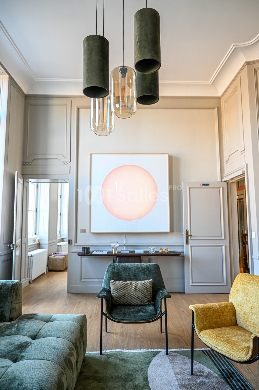 Salon lumineux avec fauteuils colorés, suspension moderne et tableau abstrait éclairé sur un mur beige.