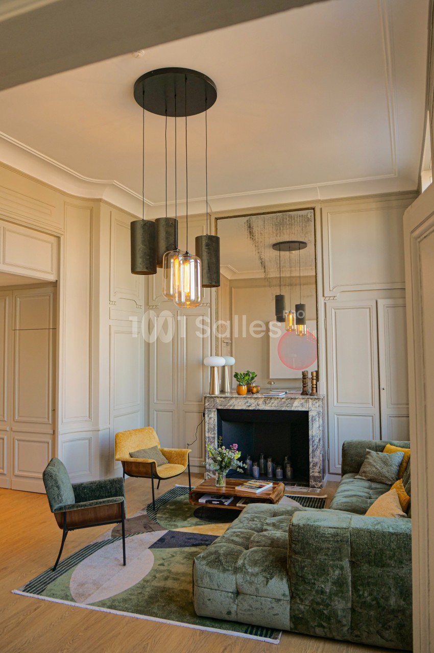 Salon élégant avec canapé vert, fauteuils jaunes, cheminée en marbre, miroir, et suspension lumineuse moderne.