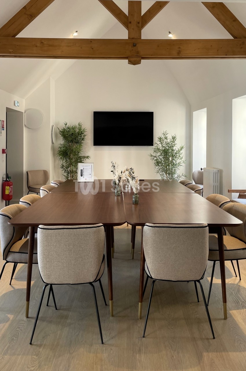 Salle de réunion lumineuse avec une grande table en bois entourée de chaises et des poutres apparentes au plafond.