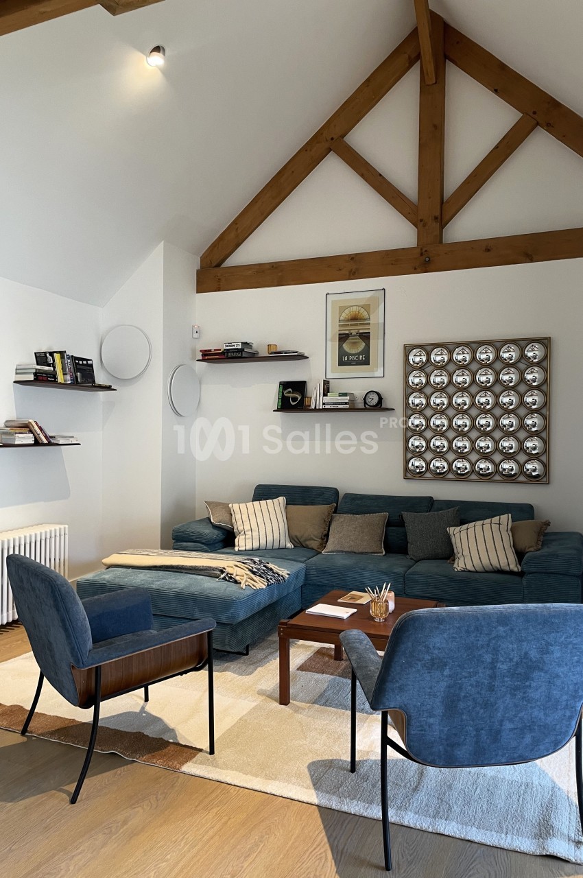 Salon moderne avec canapé bleu, fauteuils assortis, poutres apparentes, étagères murales et décoration murale géométrique.