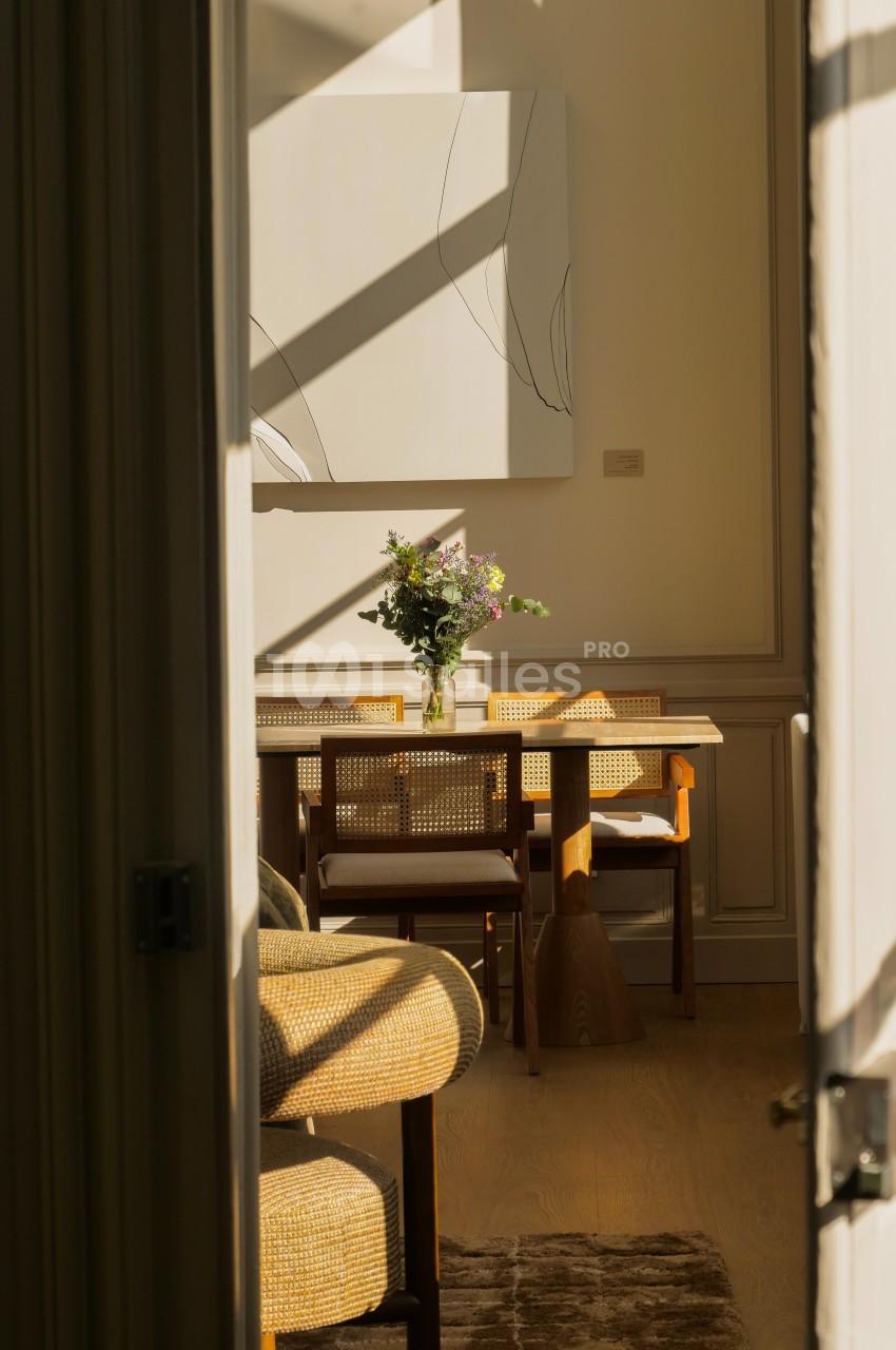 Salle à manger lumineuse avec table en bois, chaises en rotin, bouquet de fleurs et tableau minimaliste au mur.