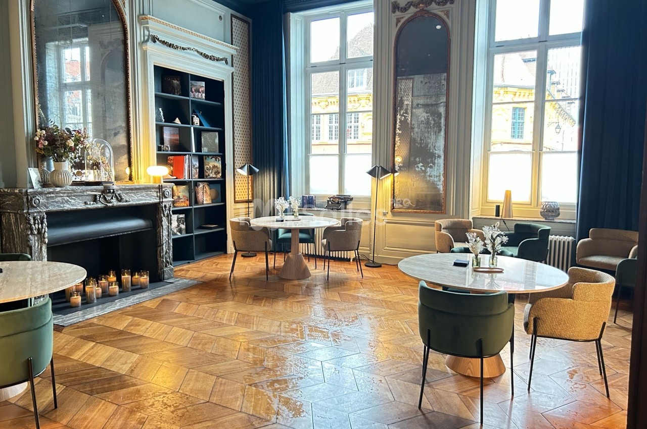 Salle élégante avec parquet en bois, tables rondes, fauteuils et cheminée décorée, éclairée par de grandes fenêtres.