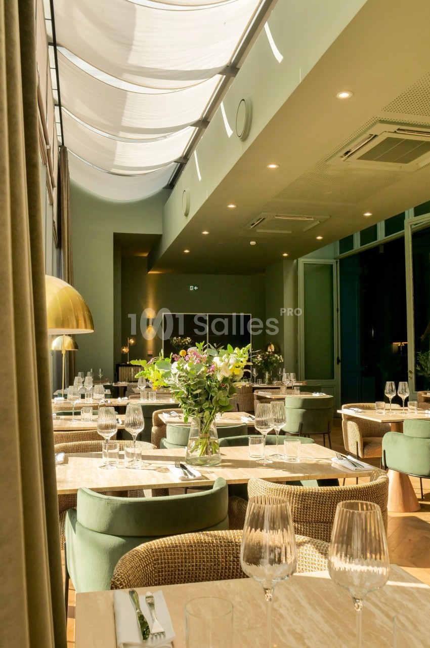 Salle de restaurant lumineuse avec tables dressées, chaises en velours vert et grande verrière laissant entrer la lumière…