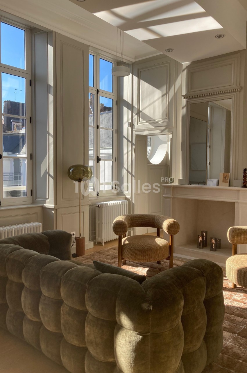 Salon lumineux avec canapé en velours, fauteuils, cheminée décorative et grandes fenêtres laissant entrer la lumière…