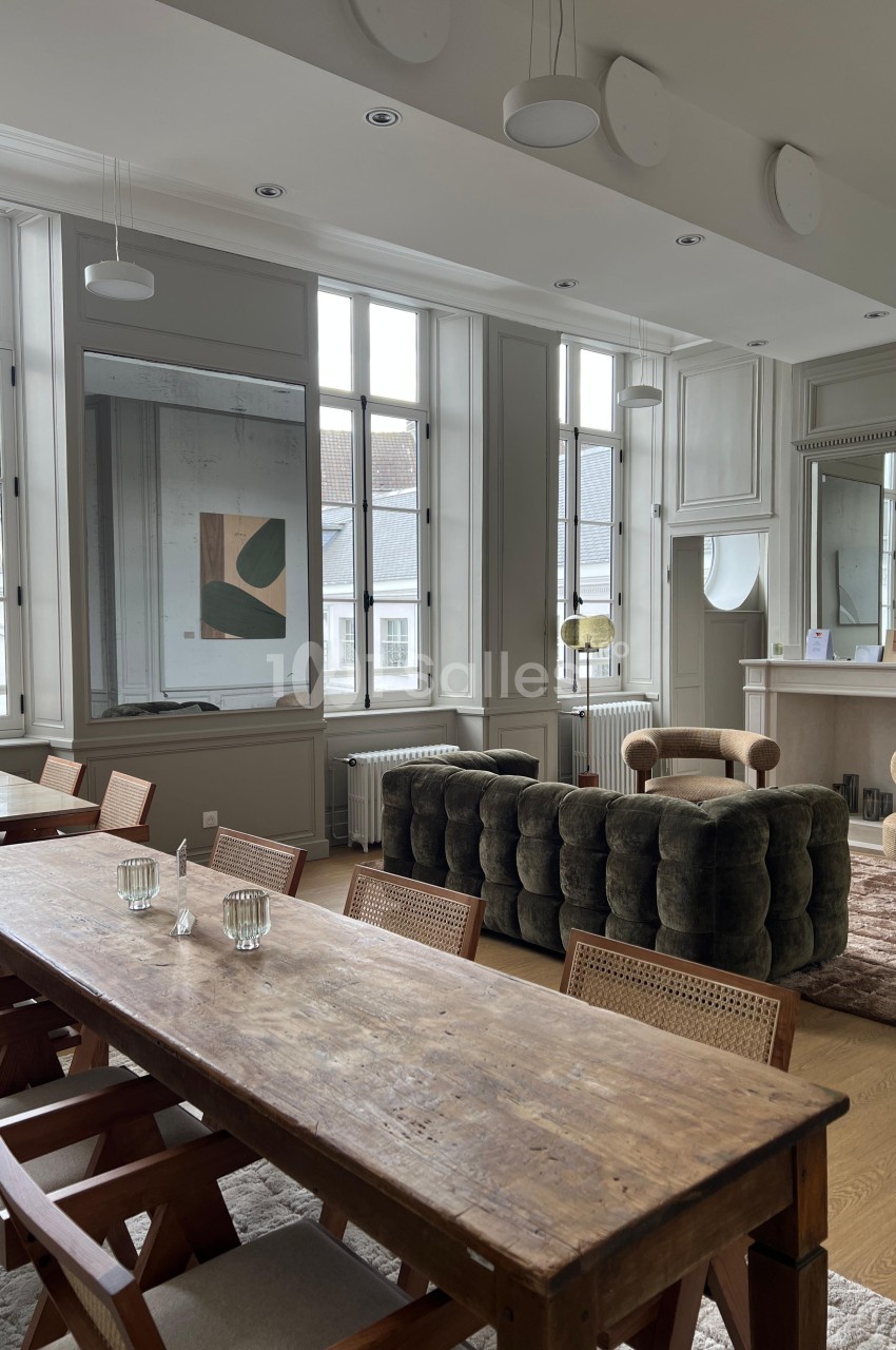 Salle lumineuse avec grandes fenêtres, table en bois, chaises, canapé vert, fauteuils beiges et décoration minimaliste.