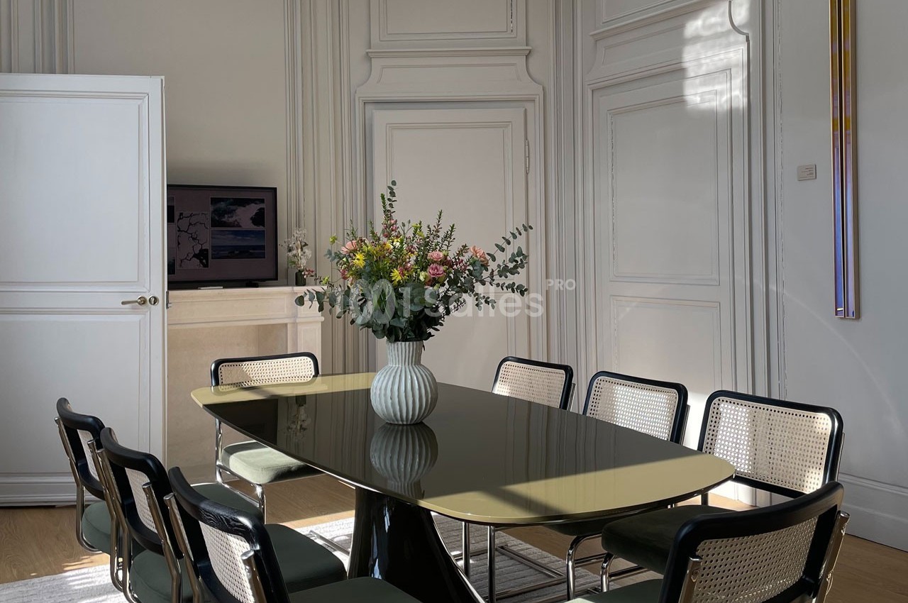 Salle à manger lumineuse avec une table ovale, des chaises en osier et un vase de fleurs colorées au centre.