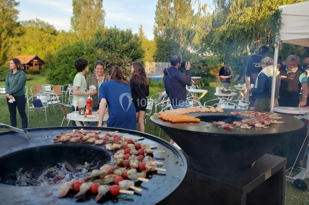 Personnes discutant près de barbecues allumés dans un jardin, avec des brochettes et saucisses en cuisson.