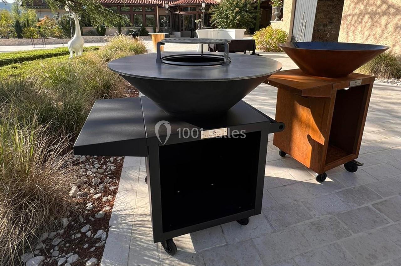 Barbecue plancha en métal noir avec tablette latérale, posé sur une terrasse en pierre, entouré de végétation.