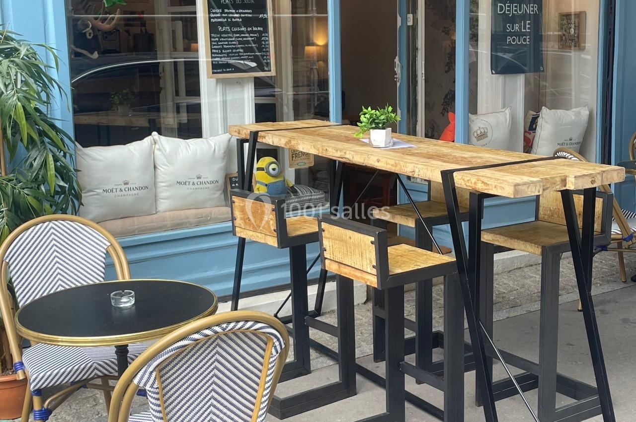 Terrasse d'un café avec tables hautes en bois, chaises en rotin et façade bleue vitrée.