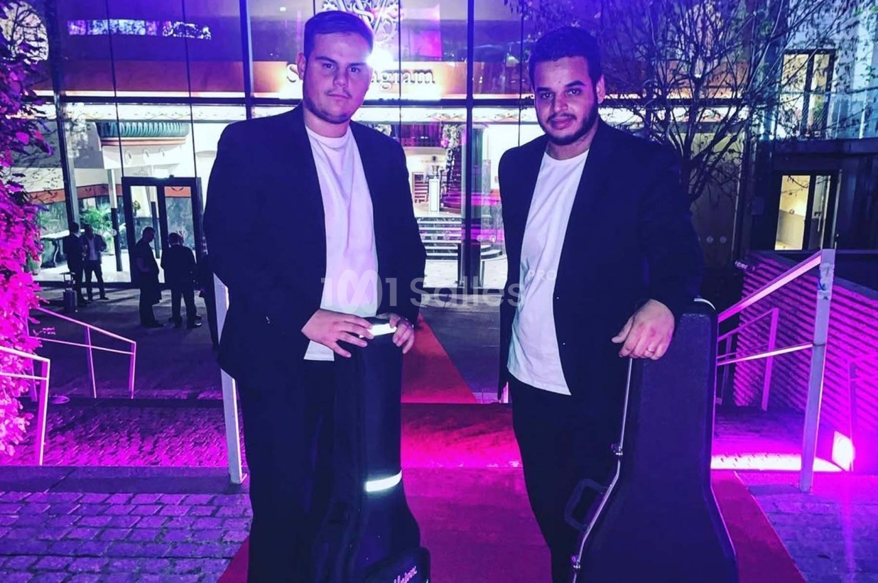 Deux hommes en costume noir tenant des étuis à guitare, debout sur un tapis rouge devant un bâtiment illuminé.