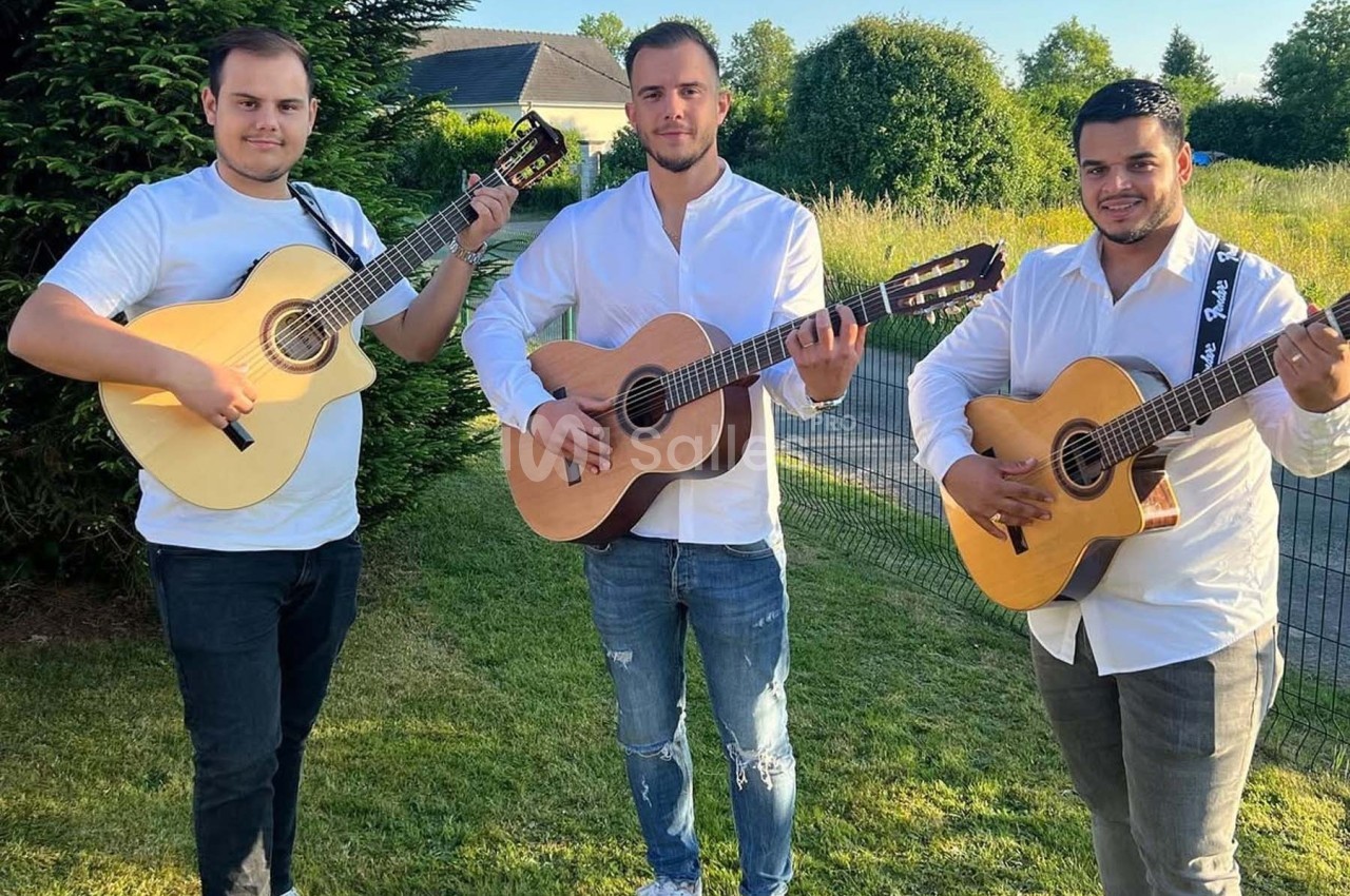 Trois hommes debout dans un jardin jouent de la guitare acoustique, vêtus de chemises blanches.
