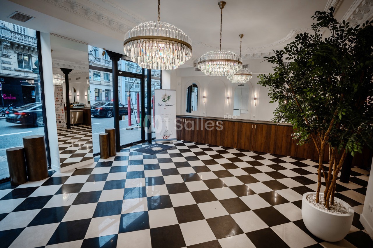 Intérieur d'un espace lumineux avec sol en damier, comptoir en bois, lustres et grandes baies vitrées donnant sur la rue.