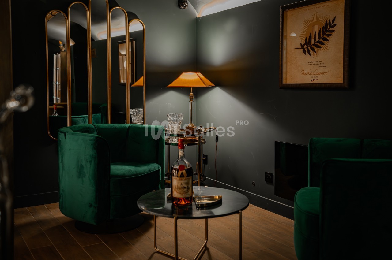 Deux fauteuils verts autour d'une table basse avec une bouteille de spiritueux, dans un salon à l'éclairage tamisé.