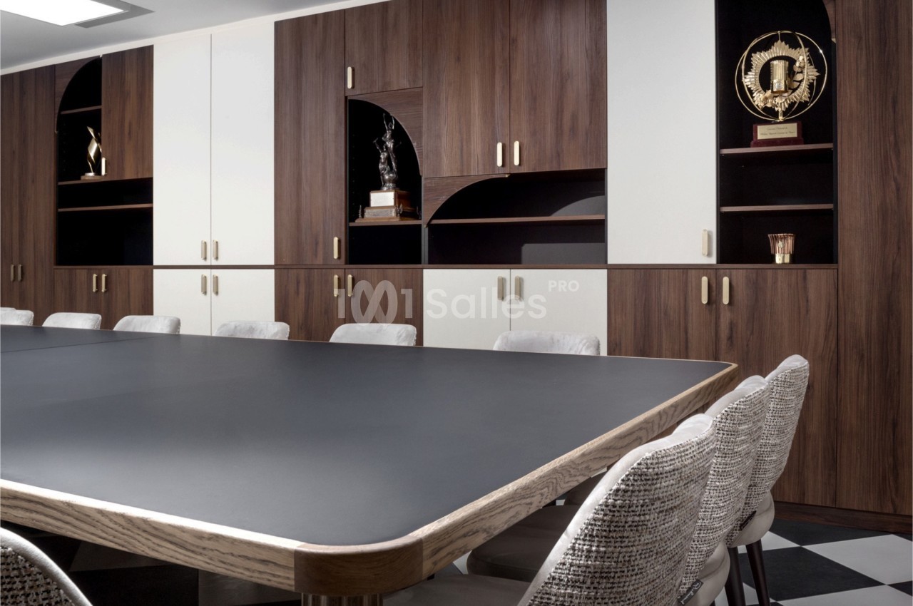 Salle de réunion avec une grande table noire, des chaises en tissu clair et des étagères en bois contenant des trophées.