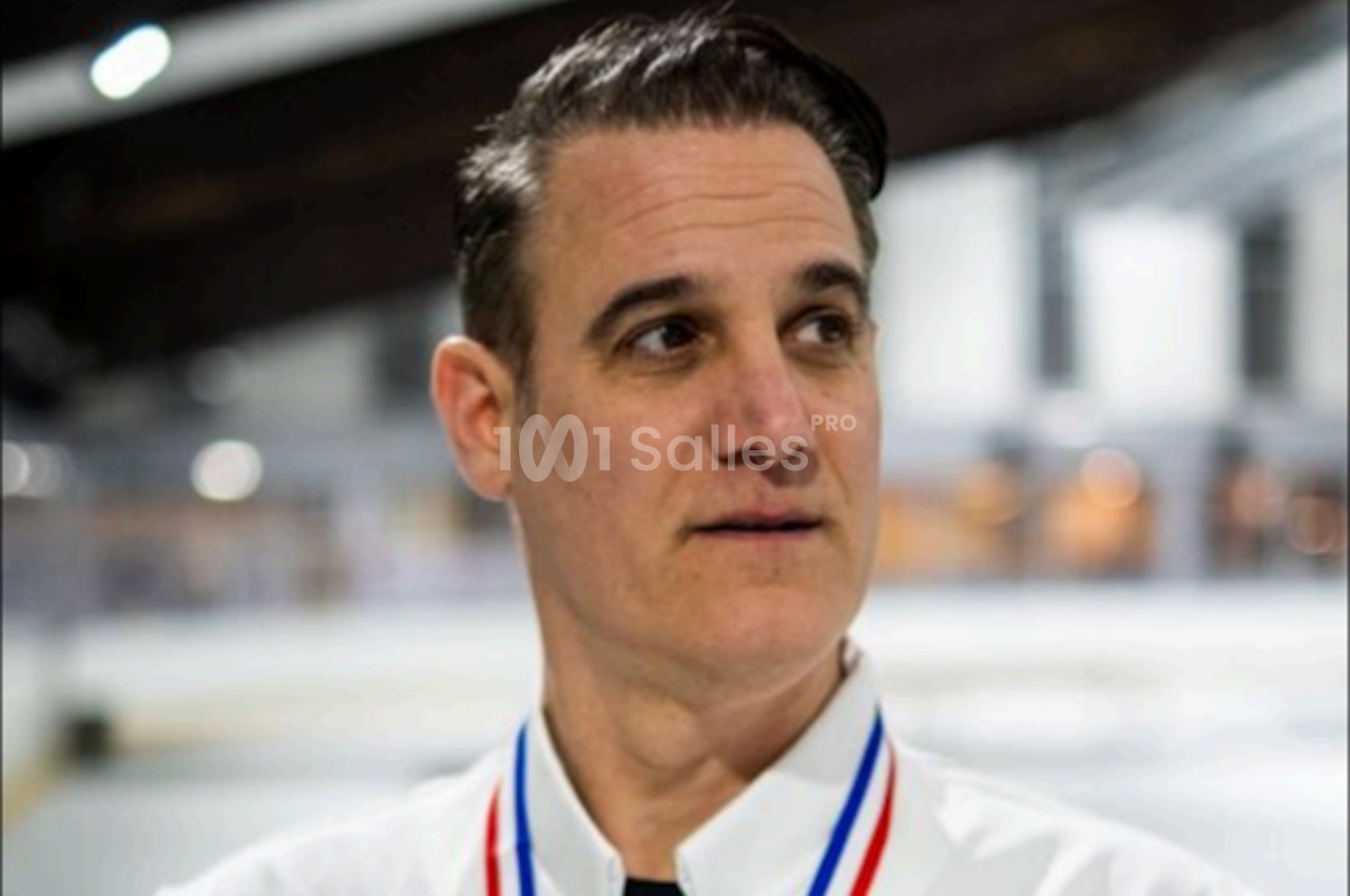 Homme portant une chemise blanche et une médaille, debout dans un environnement intérieur lumineux flou.
