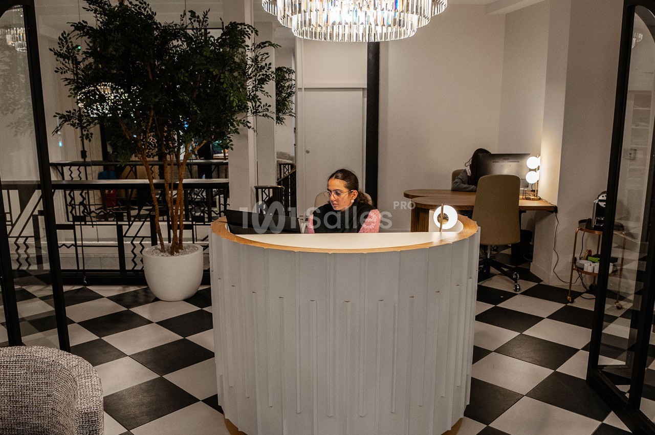Femme assise à un comptoir d'accueil arrondi dans un espace intérieur moderne avec sol en damier et éclairage suspendu.