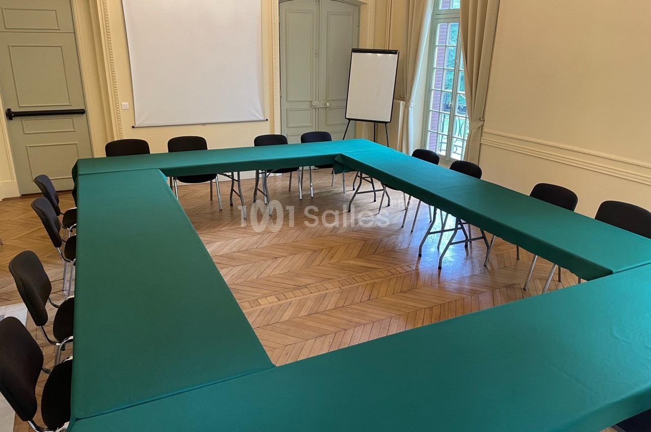 Salle de réunion avec tables disposées en U, nappes vertes, chaises noires et un tableau blanc en arrière-plan.