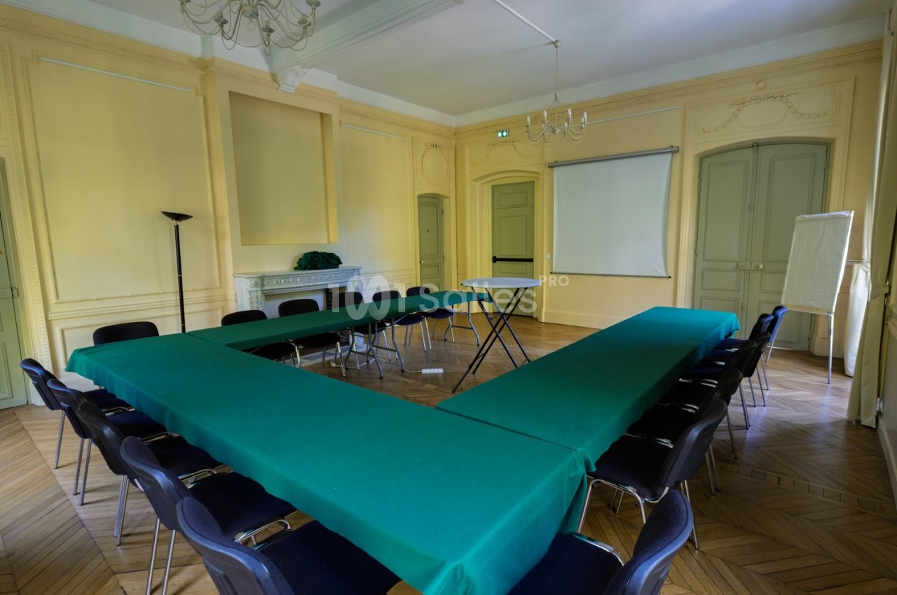 Salle de réunion avec tables en U recouvertes de nappes vertes, chaises noires, écran de projection et tableau de conférence.