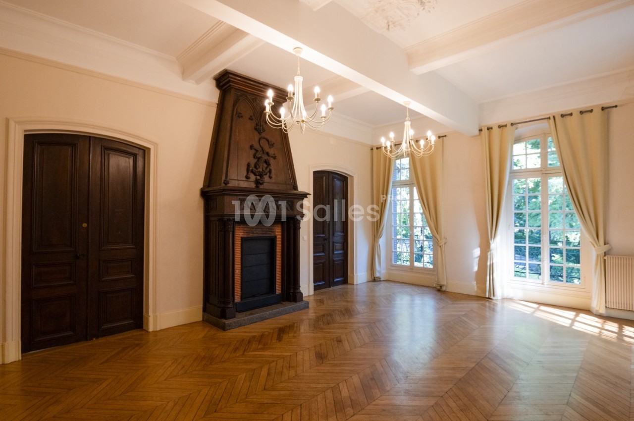 Salon lumineux avec parquet en chevrons, cheminée en bois sculpté, lustres et grandes fenêtres avec rideaux clairs.
