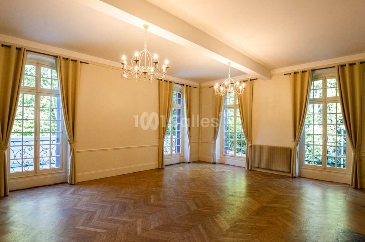 Salle lumineuse avec parquet en chevrons, grandes fenêtres avec rideaux beiges et lustres suspendus.