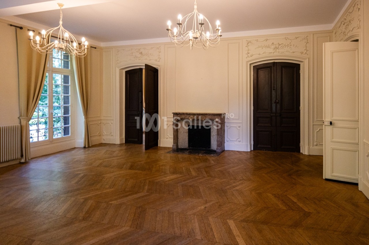 Salle vide avec parquet en bois, cheminée en pierre, murs ornés de moulures et grandes fenêtres avec rideaux beiges.