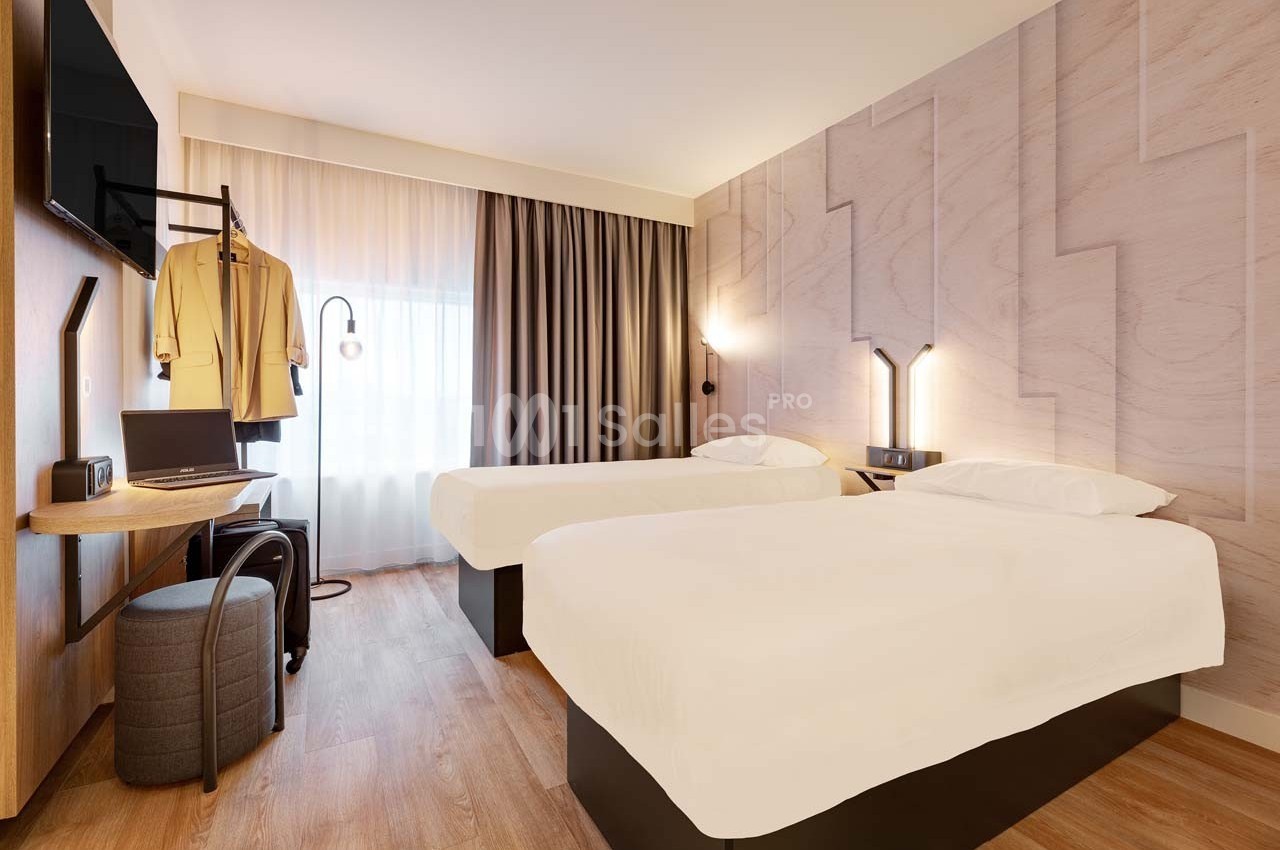 Chambre d'hôtel moderne avec deux lits simples, bureau, télévision murale et grande fenêtre avec rideaux.