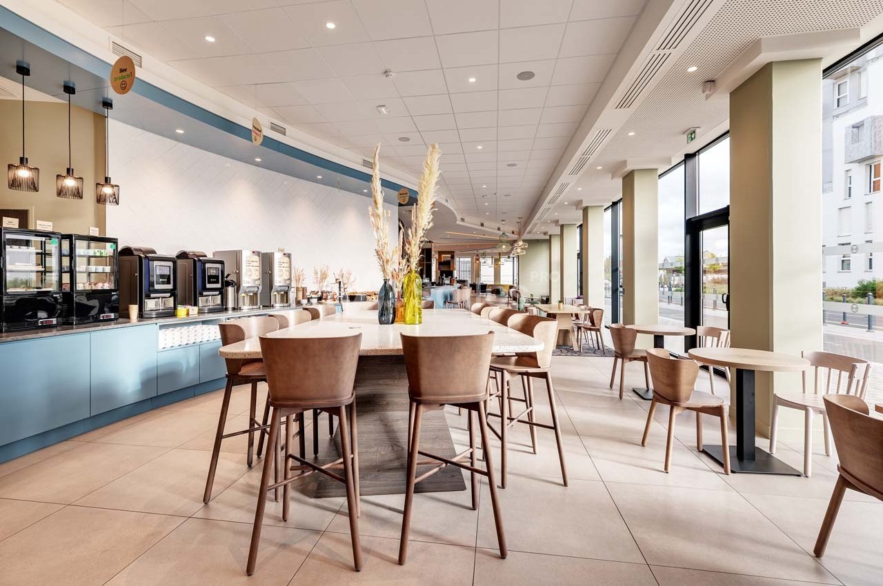 Salle lumineuse avec tables et chaises en bois, comptoir de service à gauche et grandes baies vitrées à droite.