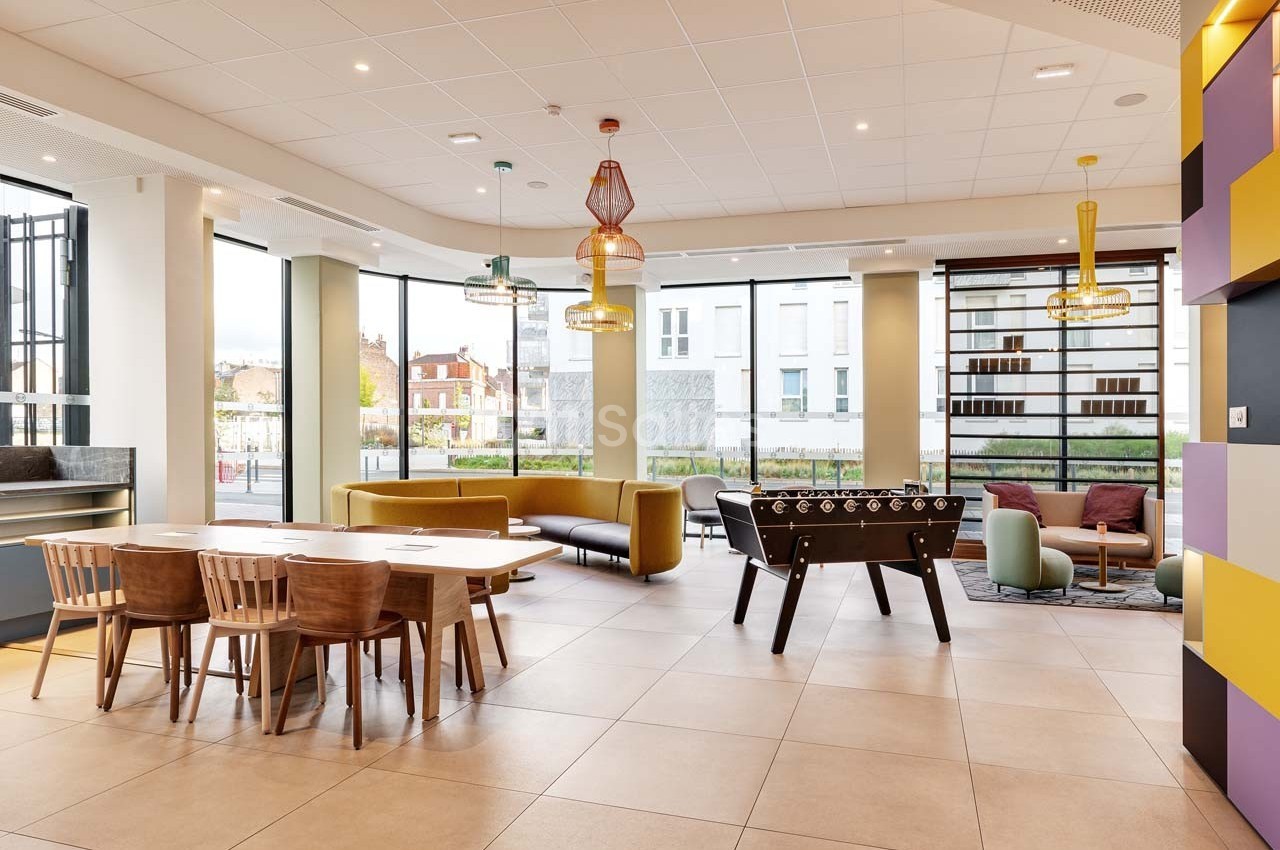 Espace lumineux avec tables en bois, canapés colorés, baby-foot et grandes baies vitrées donnant sur l'extérieur.