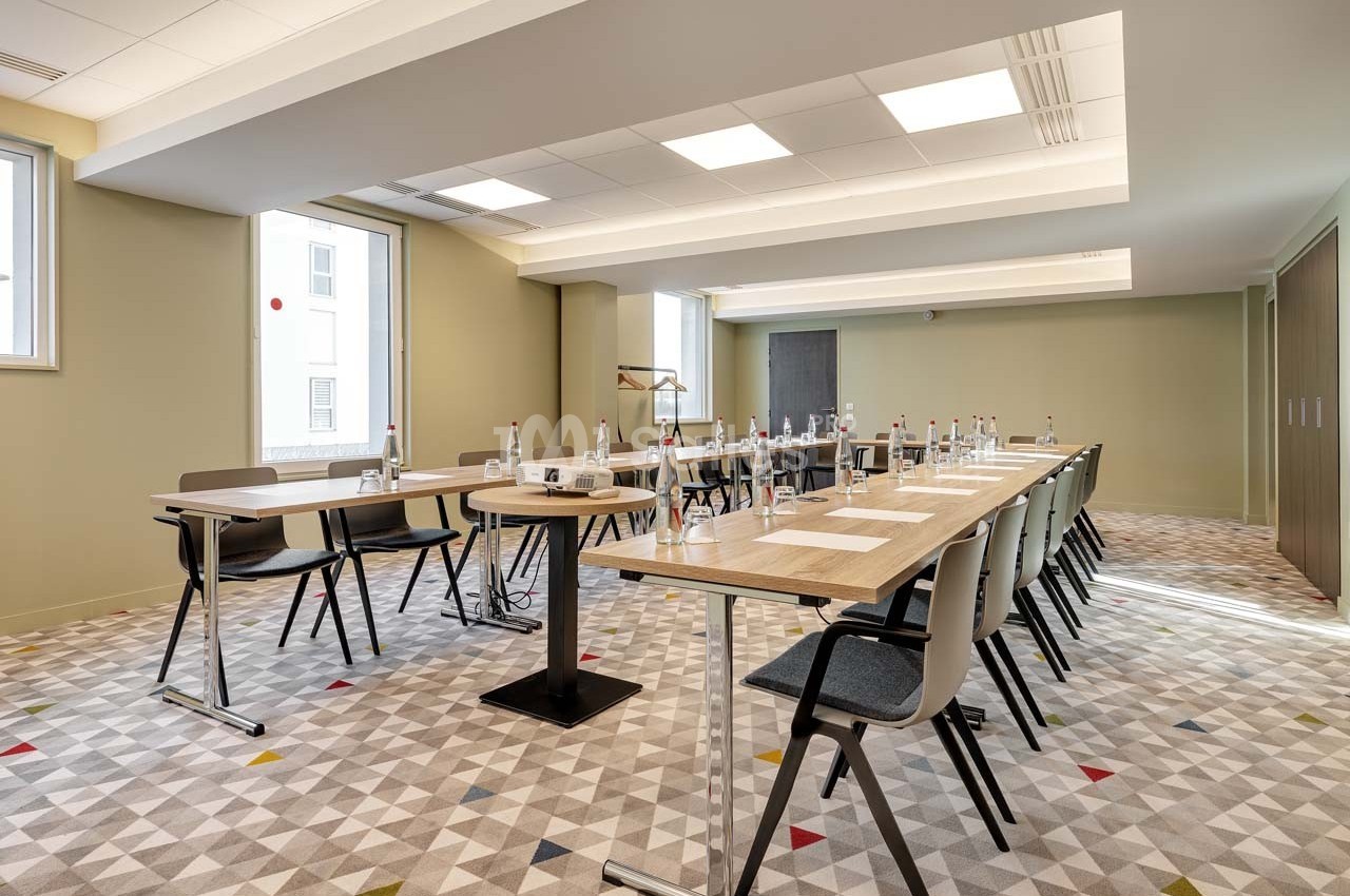 Salle de réunion lumineuse avec tables alignées, chaises modernes, bouteilles d'eau et matériel de présentation.