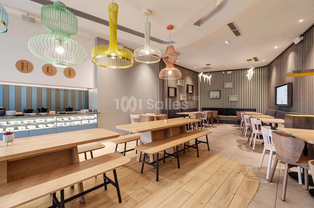 Salle de restaurant moderne avec tables en bois, chaises variées et luminaires colorés suspendus au plafond.