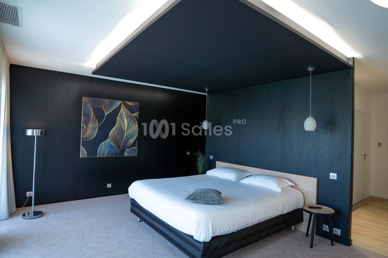 Chambre moderne avec lit double, murs sombres, tableau décoratif, lampes suspendues et grande fenêtre à gauche.