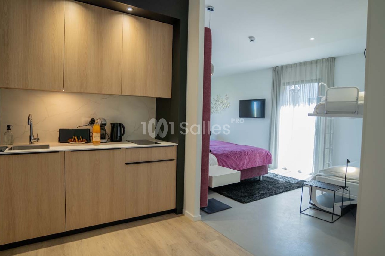 Cuisine moderne avec placards en bois, ouverte sur une chambre avec lit double, lit superposé et télévision murale.
