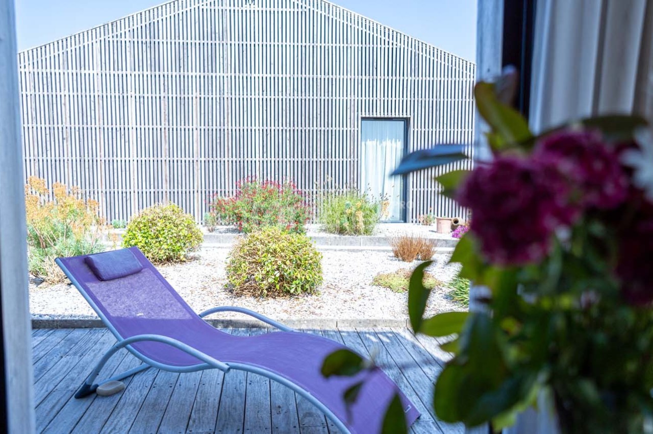 Chaise longue violette sur une terrasse en bois, avec vue sur un jardin et un bâtiment métallique en arrière-plan.
