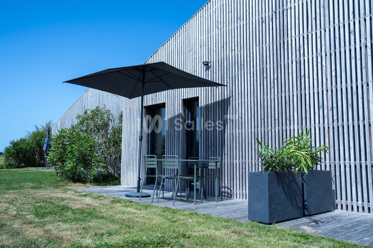 Terrasse en bois avec table, chaises et parasol, entourée de plantes, devant une façade en bois sous un ciel bleu.
