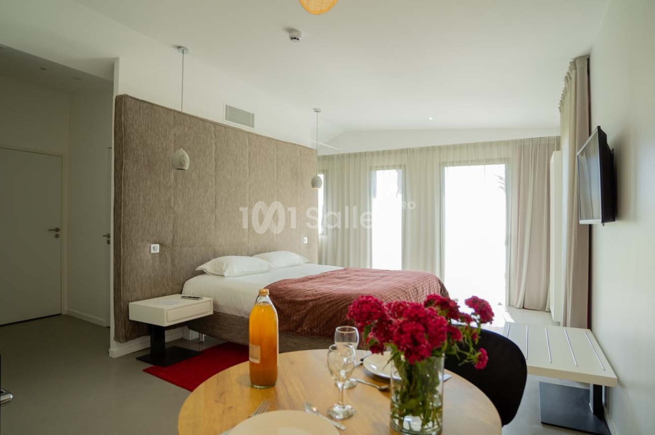 Chambre lumineuse avec lit double, table dressée, fleurs rouges, télévision murale et grandes fenêtres avec rideaux.