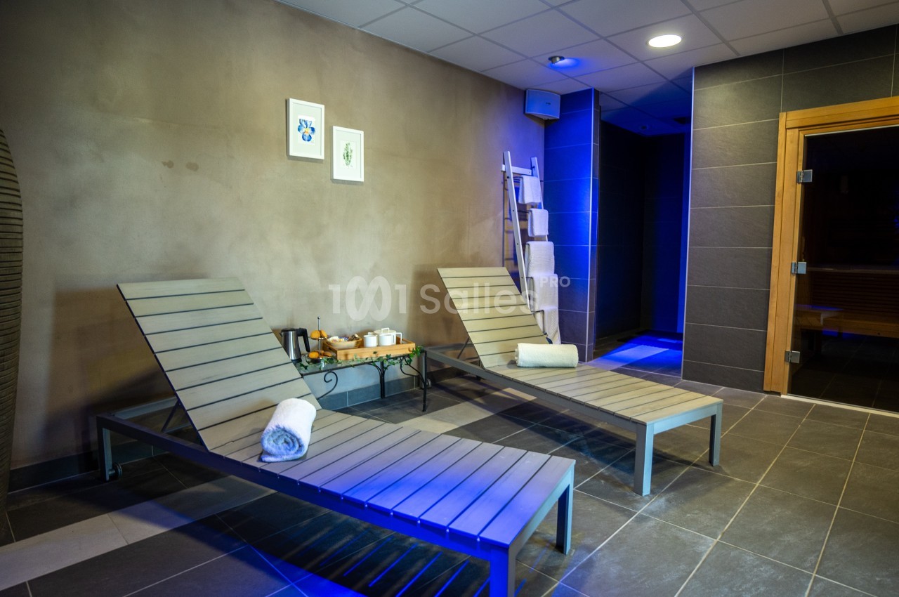 Deux chaises longues en métal avec serviettes roulées dans un espace spa, près d'un sauna en bois.