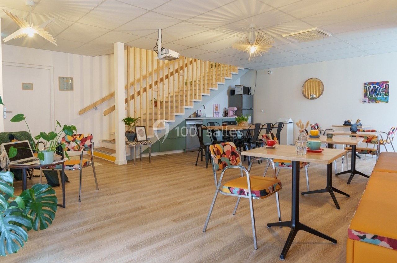 Salle lumineuse avec tables et chaises colorées, escalier en bois, plantes décoratives et ambiance conviviale.