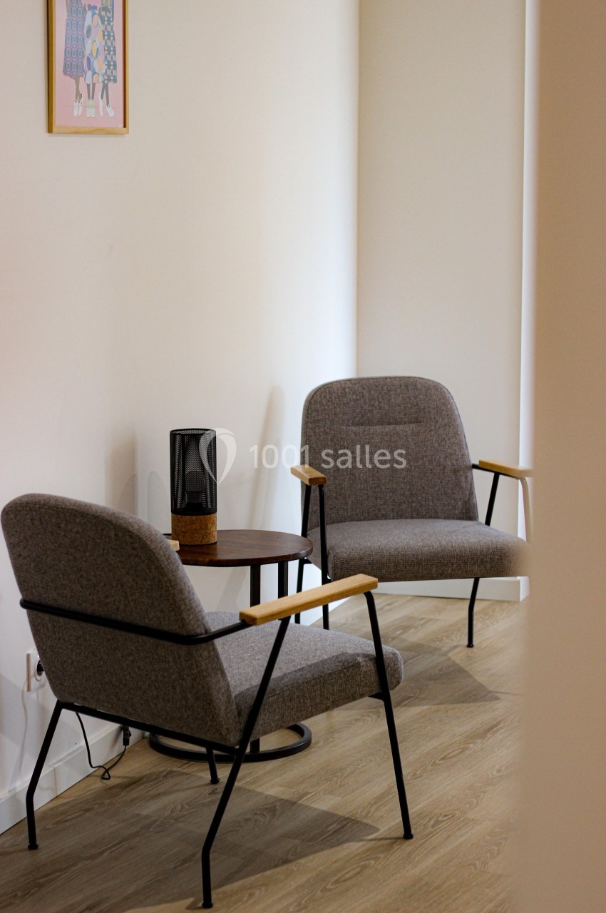 Deux fauteuils gris avec accoudoirs en bois entourent une petite table ronde dans une pièce lumineuse.