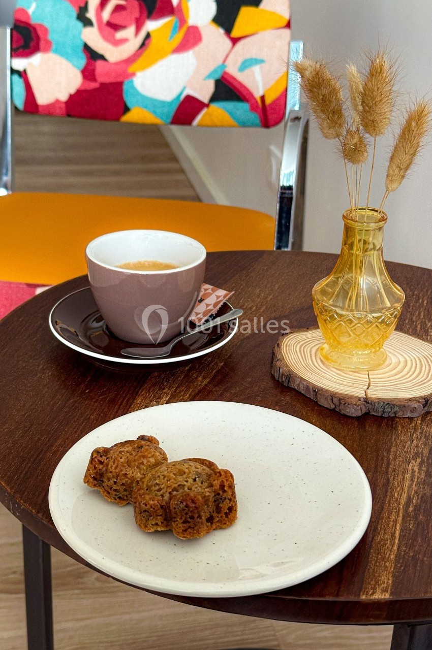 Tasse de café et assiette avec des gâteaux sur une table en bois, décorée d'un vase et d'une chaise colorée en arrière-plan.