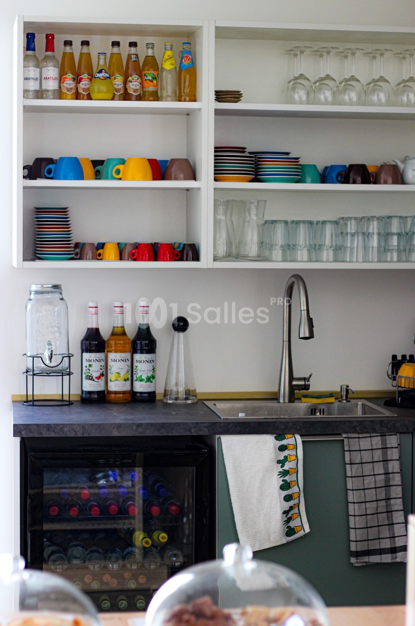 Cuisine avec étagères ouvertes contenant des verres, tasses colorées, bouteilles et un évier en inox à côté d'un mini-frigo.