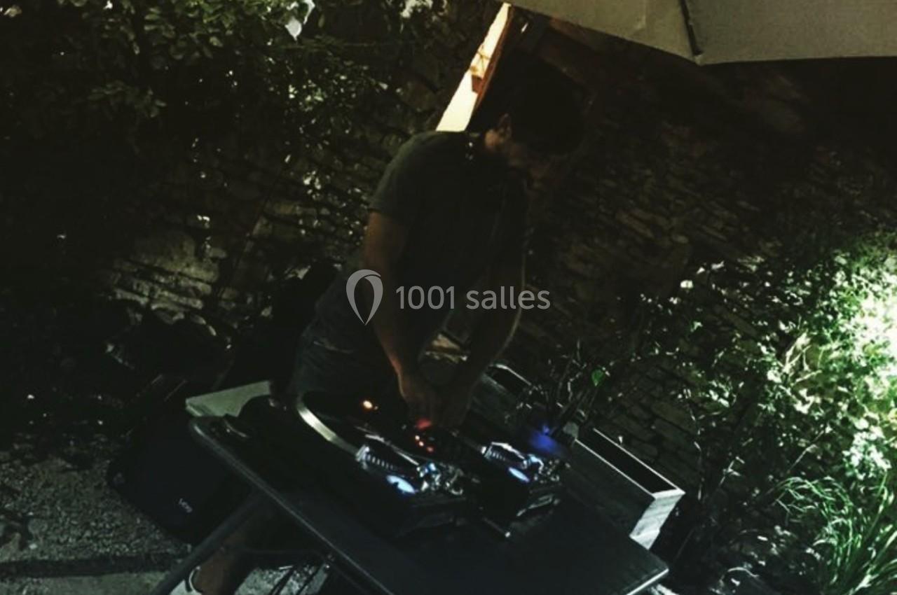 Un DJ mixe sur une table de mixage en extérieur, sous un parasol, entouré de végétation et de lumière tamisée.