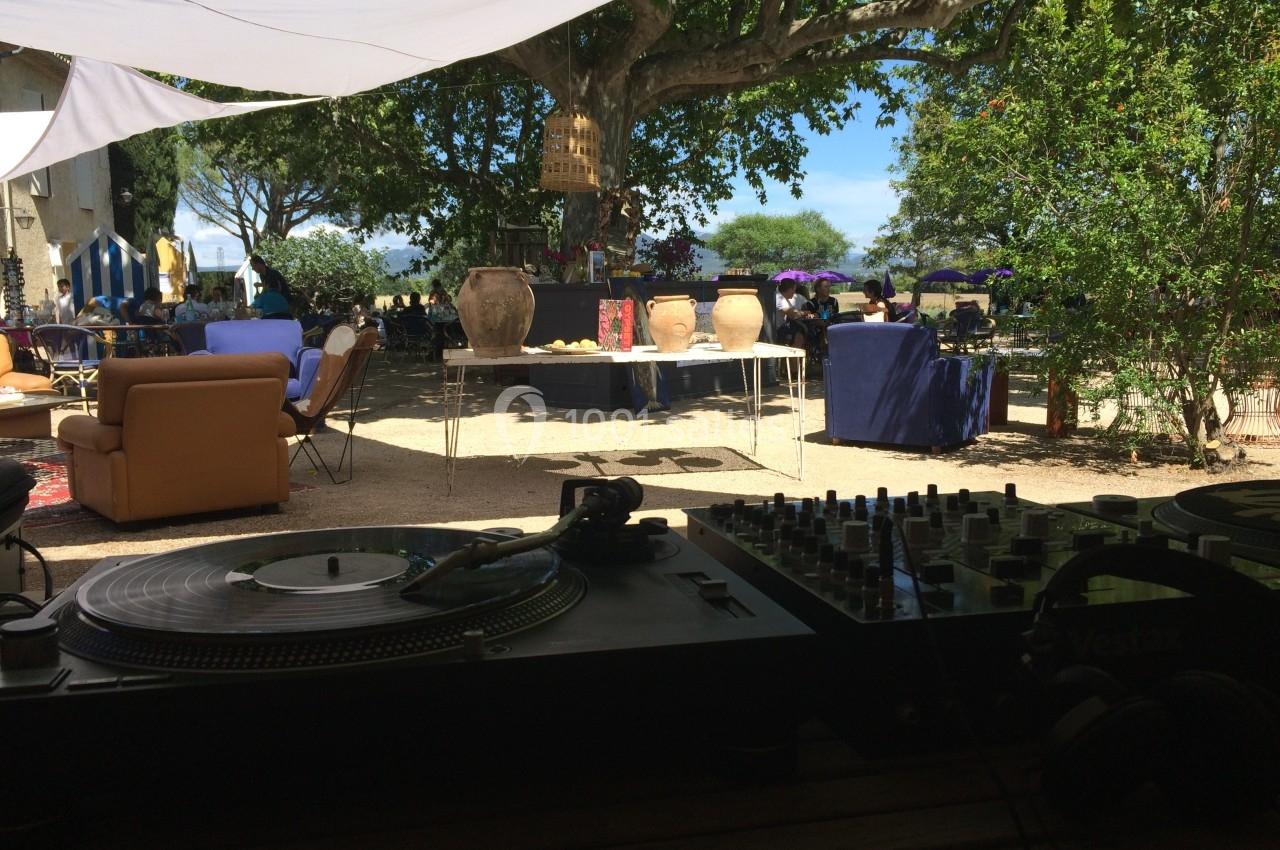 Vue depuis une table de mixage sur une terrasse ombragée avec des fauteuils, des jarres et des stands en arrière-plan.