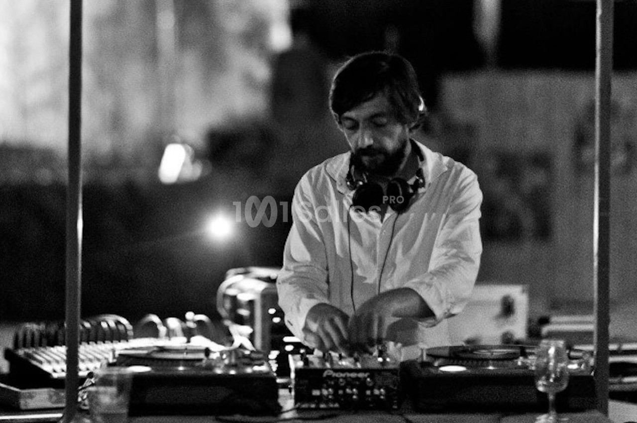 Un DJ en chemise blanche mixe de la musique sur une table de mixage dans un environnement faiblement éclairé.