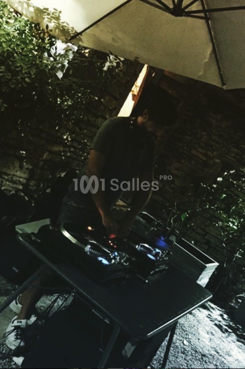 Un DJ mixe sur une table de mixage sous un parasol dans un jardin verdoyant, en soirée.