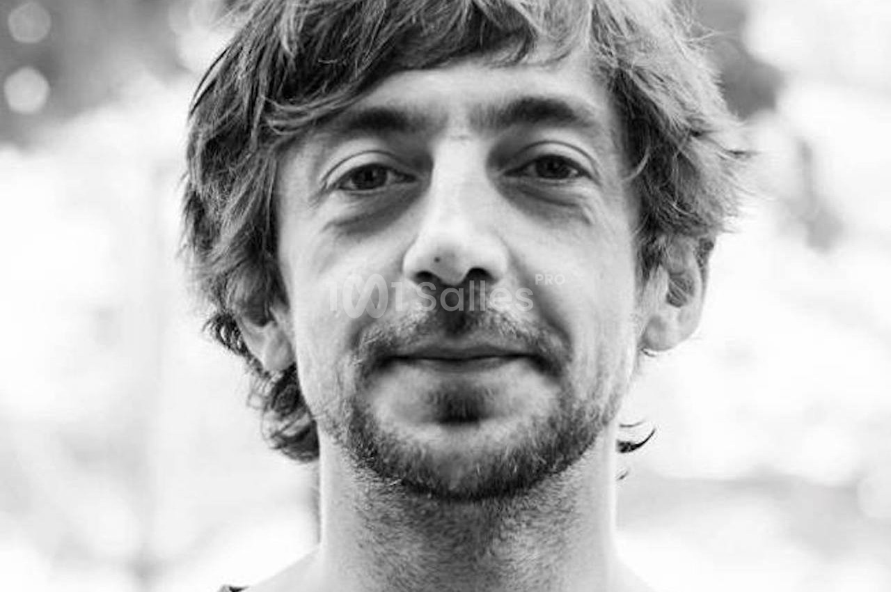 Portrait en noir et blanc d'un homme barbu avec des cheveux mi-longs, portant un t-shirt, en extérieur flou.