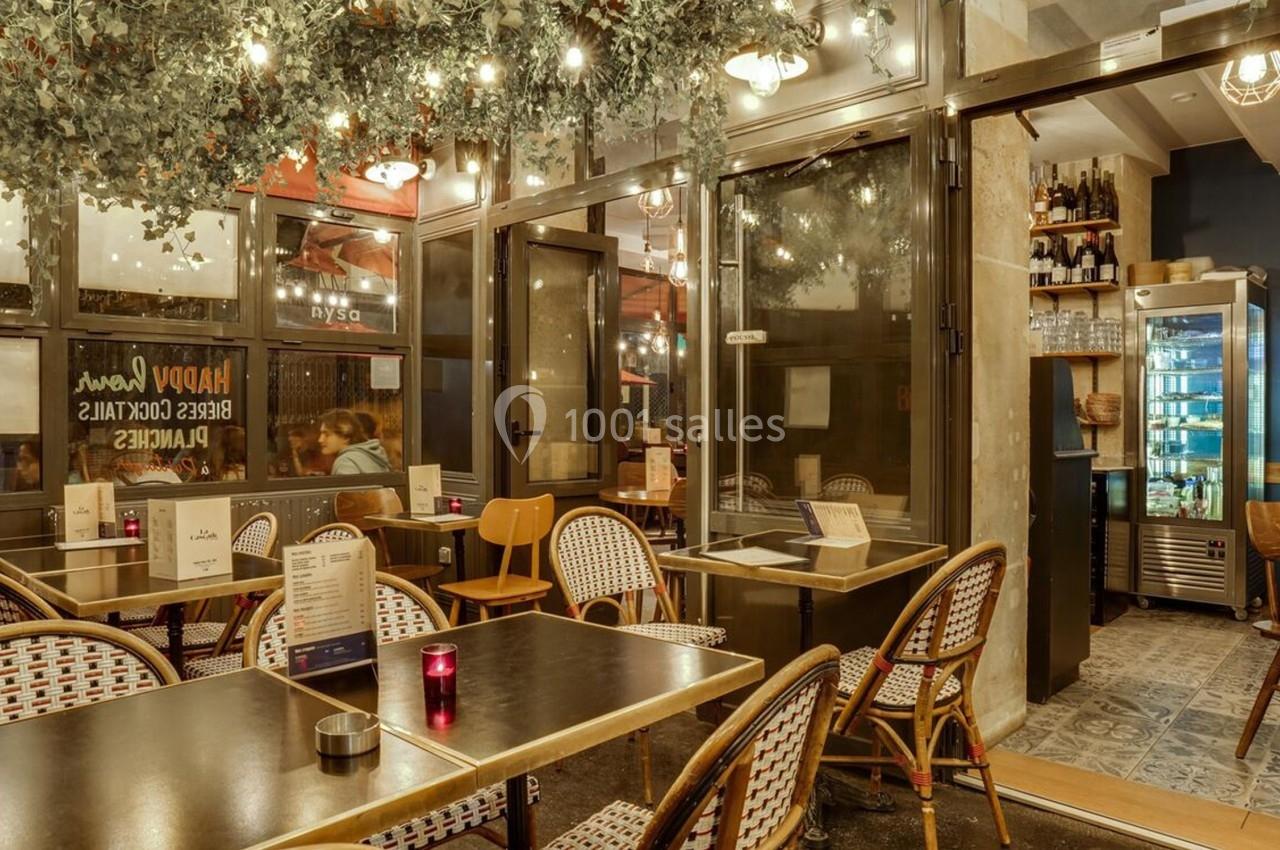 Salle de restaurant avec tables et chaises en rotin, éclairage chaleureux et décoration végétale suspendue.